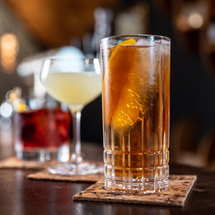 Row of three cocktails on a bar. From front to back, a long drink, a spritzer cocktail and a lowball cocktail